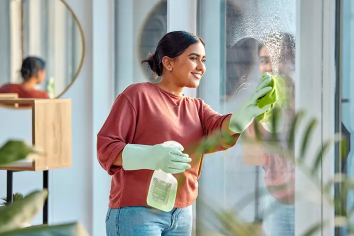 A woman cleaning a window with a green cloth while wearing gloves and holding window cleaner in one hand.