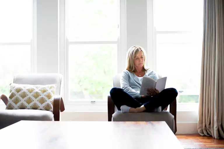 Woman reading a book in front of three large double-hung windows.