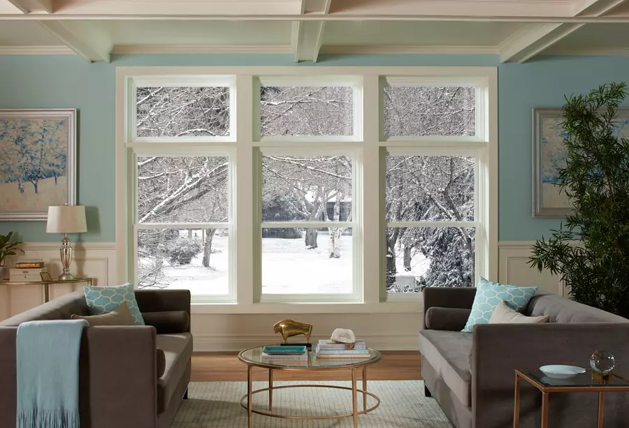 Picture of a light blue living room in the winter with picture windows displaying snow-covered ground and trees. 