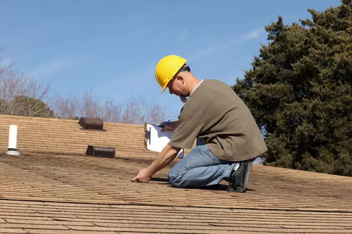 A worker wearing a hard hat inspects a residential roof while consulting a roof maintenance checklist.