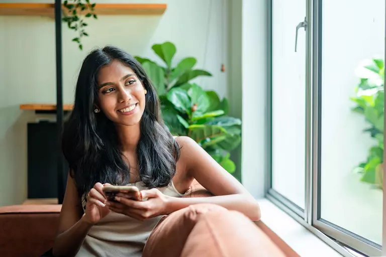 Young woman relaxing on a sofa with smartphone in hand and looking away with a smile.