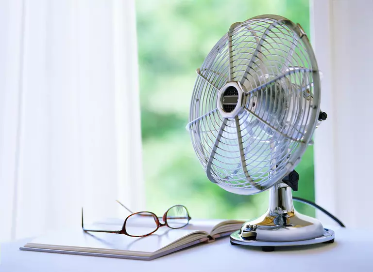 Simple, small house fan on a table next to an open book and reading glasses.
