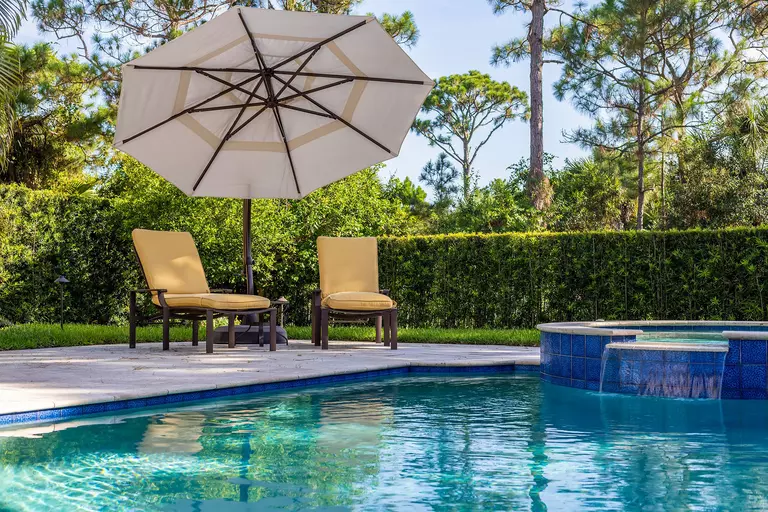 A back patio leading to a pool, surrounded by lounge chairs, an umbrella, and lots of landscaping to enclose the space.