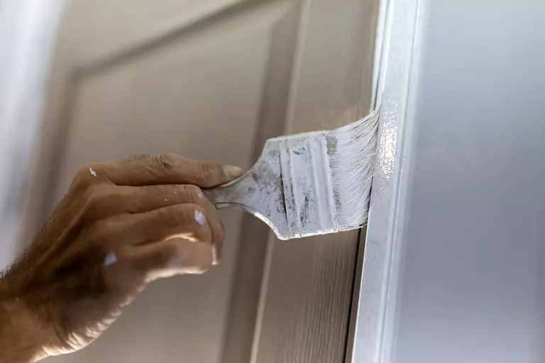 A person’s hand using a brush to apply white paint to the side of a door frame.