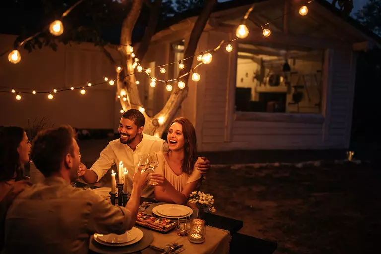 Two couples having dinner in their outdoor dining area with string lights hung up in the yard to illuminate the meal.