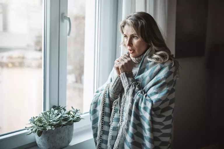 A woman in multiple layers shivering in front of a drafty window.