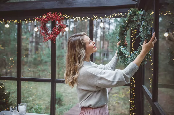 A woman hanging a wreath and string lights up on her window in a large, naturally lit
sunroom.