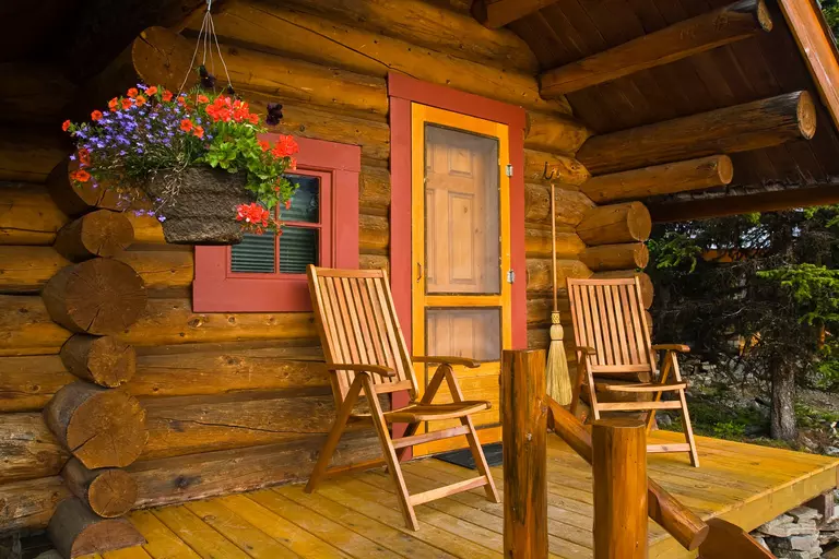 The entryway of a log cabin that has a wood-finish front door, a window with red trim, and a small front porch with an outdoor seating set. 
