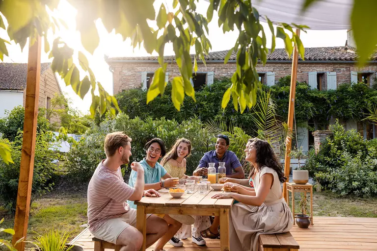 A group of friends gathered at an outdoor dining table on the back patio surrounded by landscaping and wooden décor.