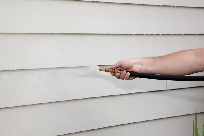 A closeup of a man’s arm while hosing the grey vinyl siding exterior of a home.