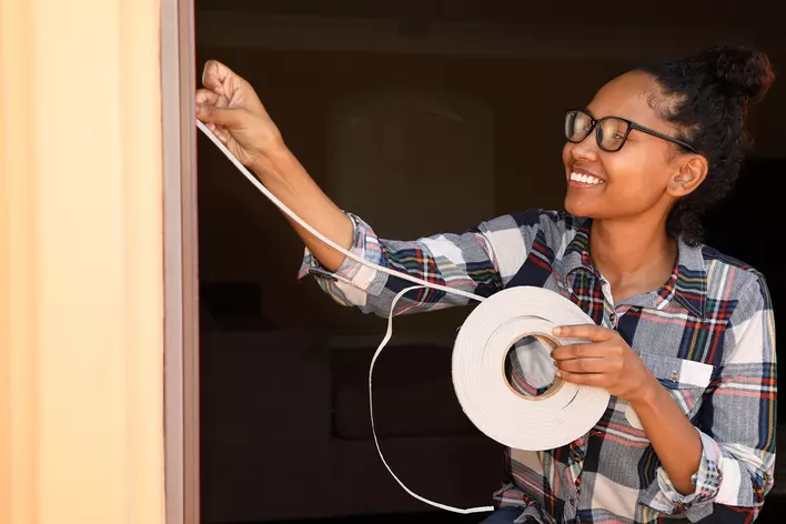 Woman smiling as she seals the leaky windows and doors of her home with
weatherstripping.