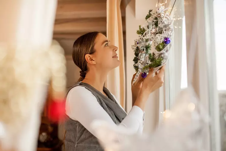 A smiling woman putting a small wreath up indoors in front of a window.
