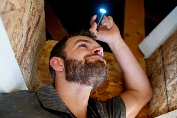 A man in the attic inspecting if there are any leaks through the roof with a flashlight.