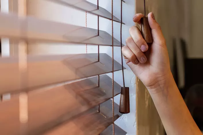 A person pulling the string of wood blinds to close them.