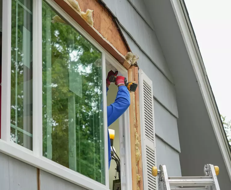 Worker inside the second story of a home using a powered drill on a window frame.