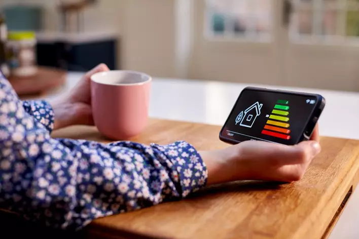 A close-up of a woman holding a smart energy meter in kitchen measuring energy efficiency with a pink mug in one hand.