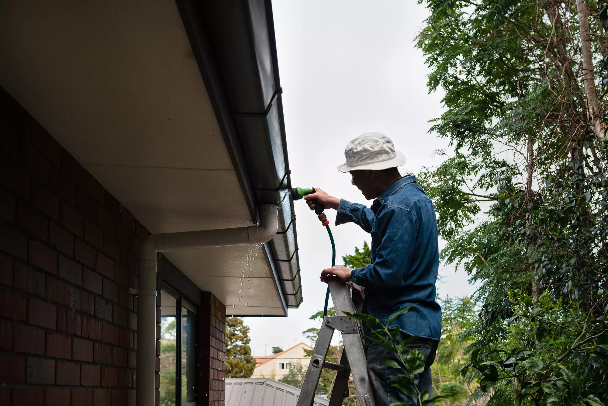 A man on a ladder rinsing out his gutters with a hose to get gunk and debris out.