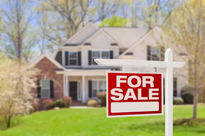 A “for sale” sign in the foreground slightly obscures the image of a two-story home in the background.