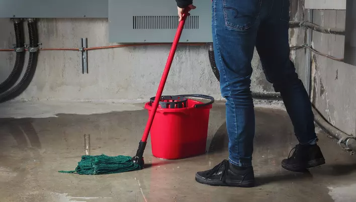 Someone mopping the floor of a concrete basement that has various pipes in it.