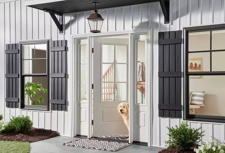 The front of a home with two double-hung windows and the front door open with a dog standing in it.