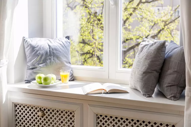 A window ledge decorated with pillows, an open book, apples, and a beverage.