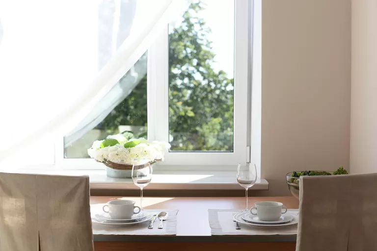 A dining table with place settings for two in front of a sunny window with a curtain partially pulled back.
