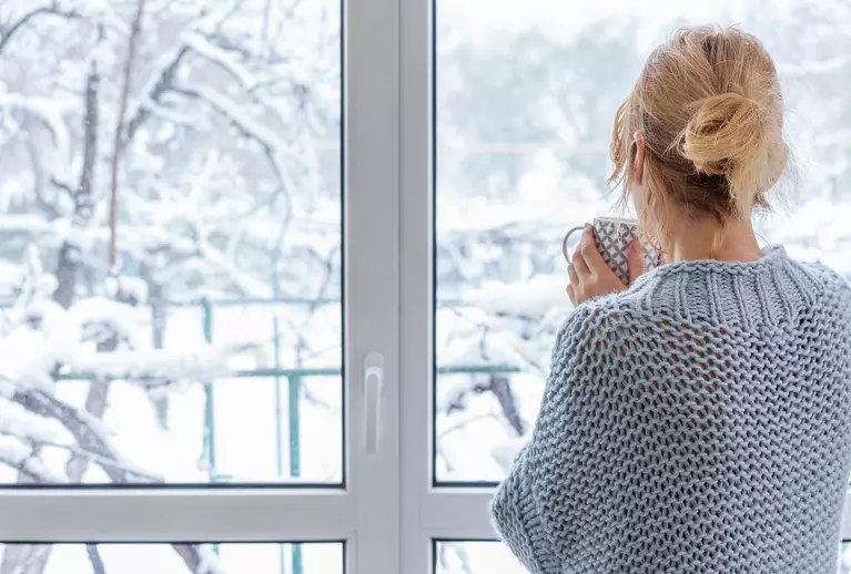 A woman with a mug looking through patio doors at the snow.