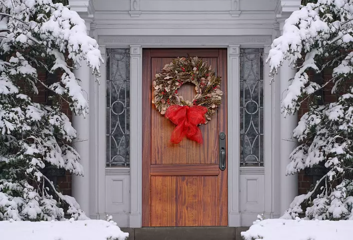 A snow-covered home with a wood grain exterior door with a Christmas wreath