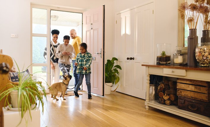 Family of four walking through the entry door of their home into the living room with a dog on a leash.