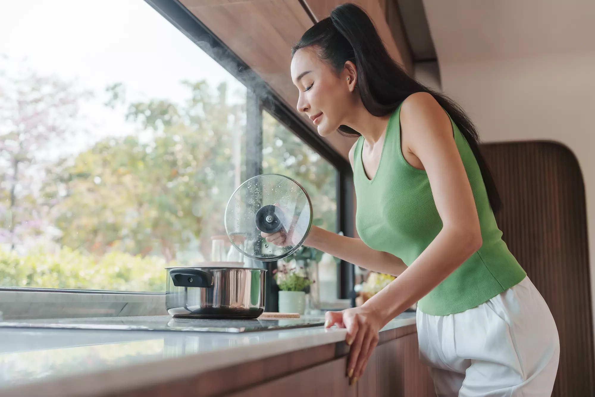 Woman cooking at stove with open kitchen window venting steam outside.