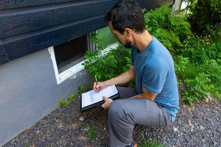 An inspector looking at the foundation of a home and a basement hopper window