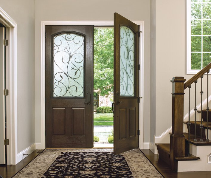 An interior view of a pair of woodgrain fiberglass doors with decorative privacy glass<br>panels opening to a sunny exterior at a home’s entrance.
