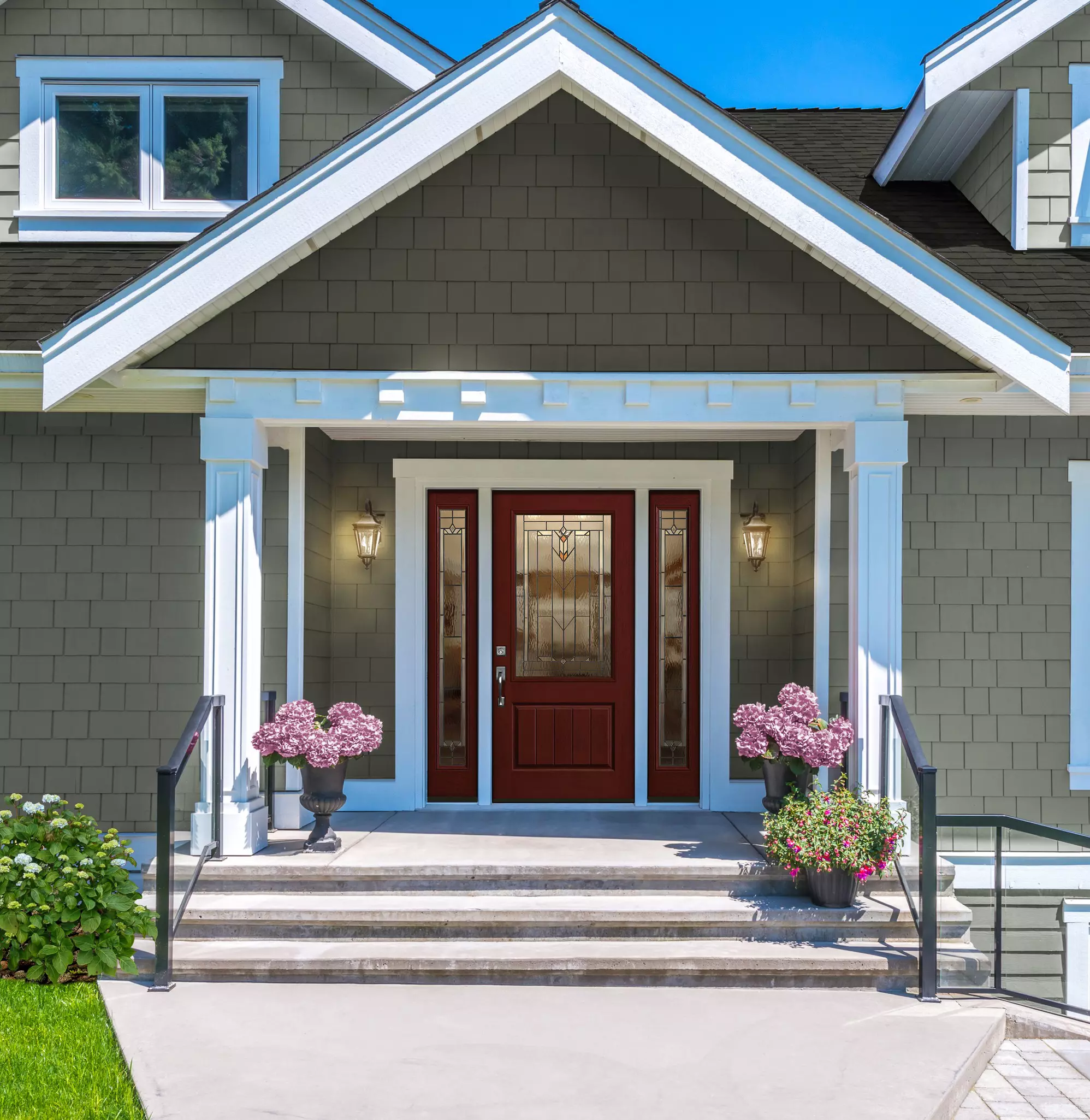The front of a sunlit gray home with an entry door flanked by blooming flowers.