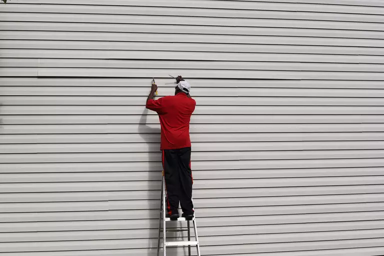 A man on a ladder repairing damaged vinyl siding on the side of a home.