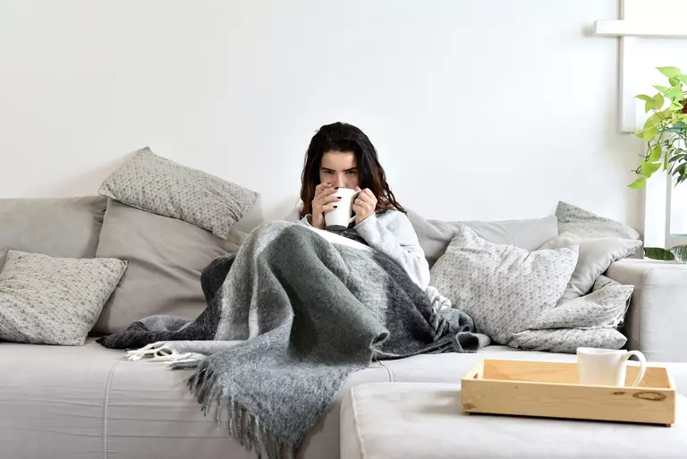 Woman sitting on a couch of a home while wrapped in a blanket, holding a mug