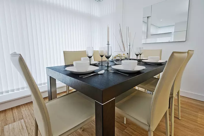 A modern dining room with light-colored walls, a wood dining table furniture set, a large window shaded with vertical blinds, and decorative dishware.
