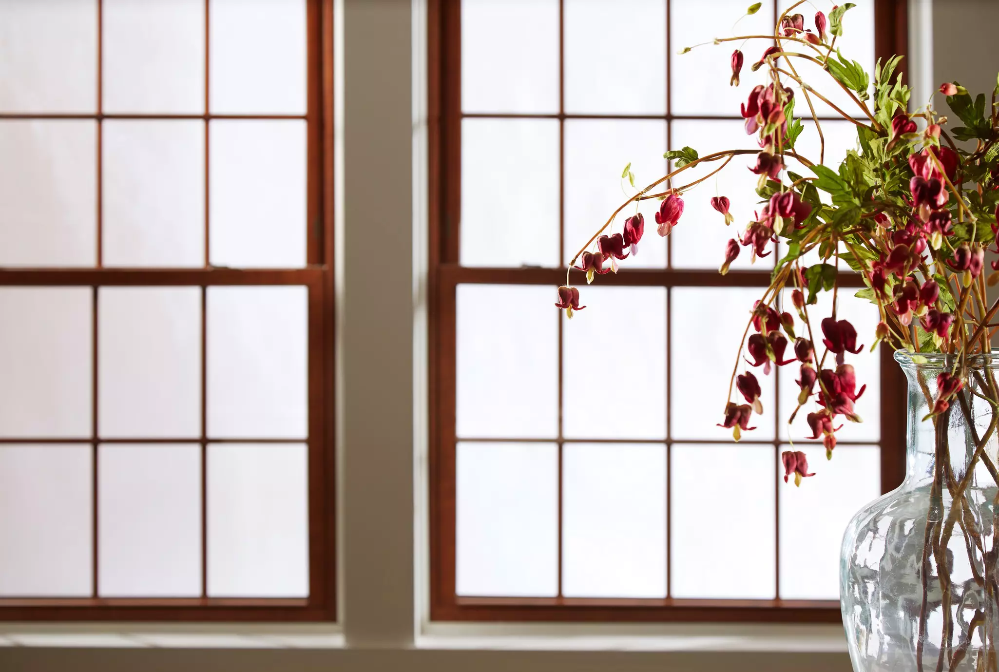 Two dark wood-framed grid windows behind a glass vase of bleeding heart flowers.