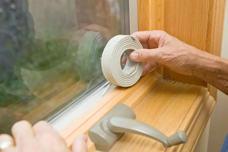 Person with a roll of weatherstripping applying the material to the edges of a window frame.