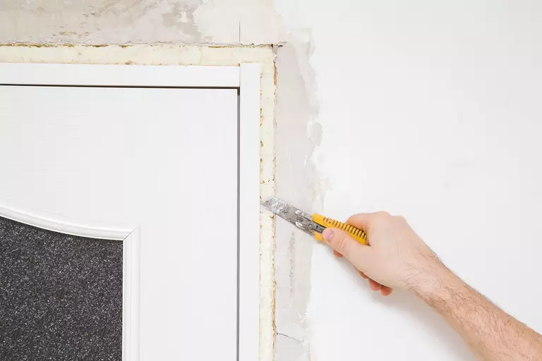 Person holding blade knife and cutting construction foam between new white wooden door and wall for exterior door installation.