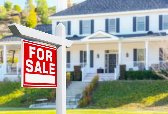 A red “For Sale” sign in front of a large white home with black exterior accents and energy-efficient windows. 