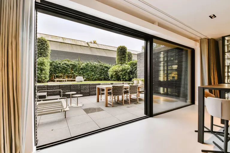 Large open sliding patio doors exiting the kitchen of a modern home that leads into a spacious and beautifully decorated back patio and yard.