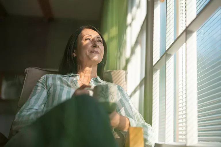 A smiling woman seated next to a window with a morning cup of coffee.