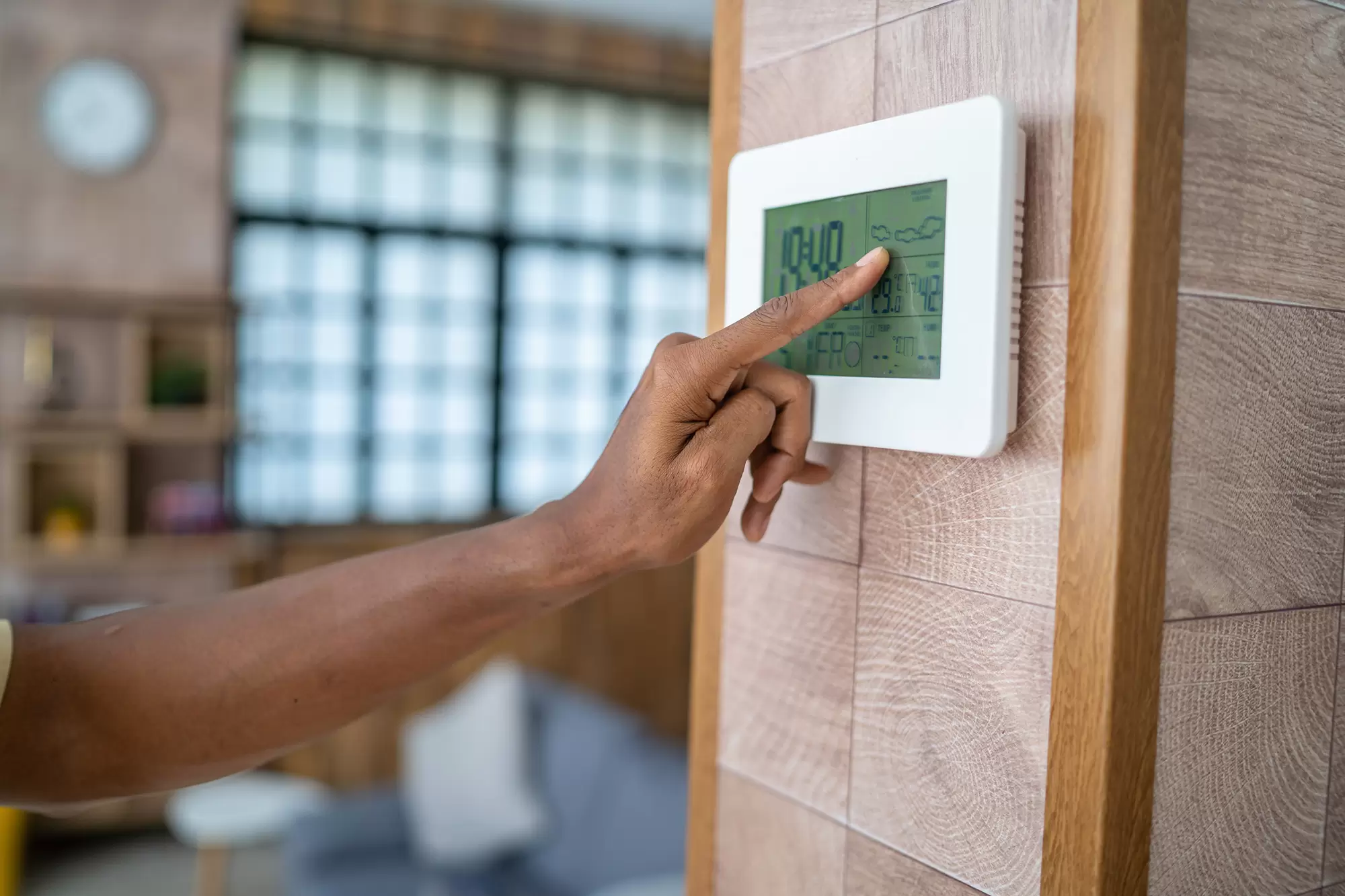 Hand adjusting digital thermostat with living room and windows in the background.