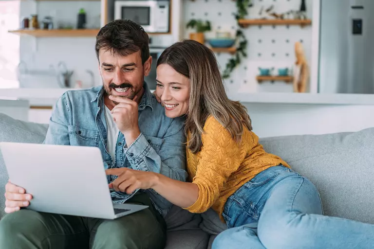 A man and a woman on a couch smiling and looking at a laptop.