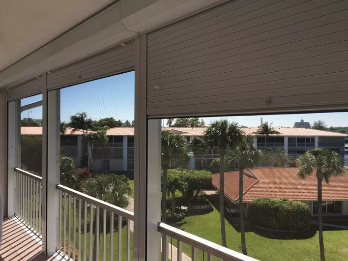 A view from inside a home that has roll-down shutters. One of the shutters is partially closed, offering a view of a sunny residential neighborhood dotted with palm trees.
