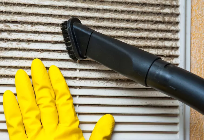 Person with yellow glove pressed up against HVAC system filter as they clean it with a vacuum brush.