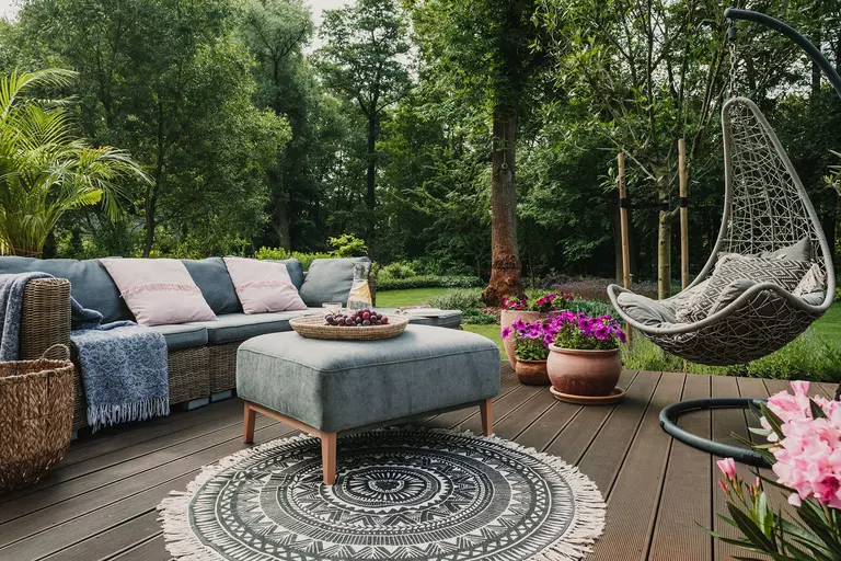 An open back patio decorated with a couch, hanging chair, ottoman, and a patterned rug to create a cozy lounge space outdoors.