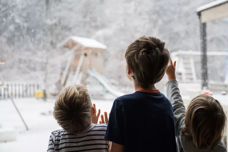 Three little kids looking out a large picture window into their snow-covered backyard.