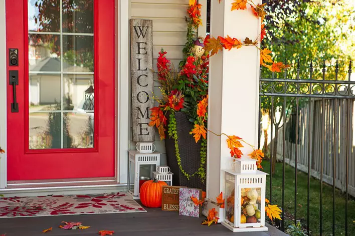 Front entrance of a home with a bright red entry door and fall décor consisting of pumpkins, dried leaves, and fall quote boards.