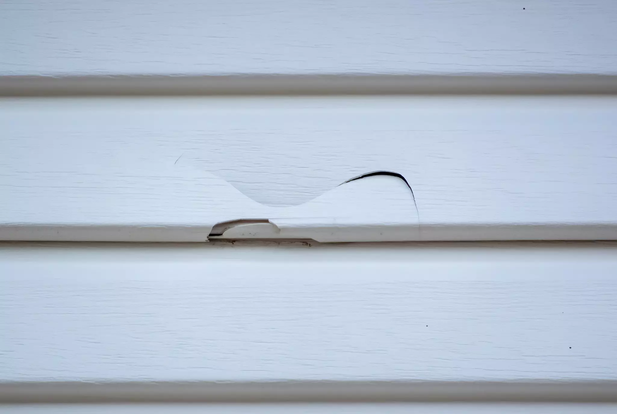 Close-up of a cracked white vinyl siding panel with visible damage and a small tear in the surface.